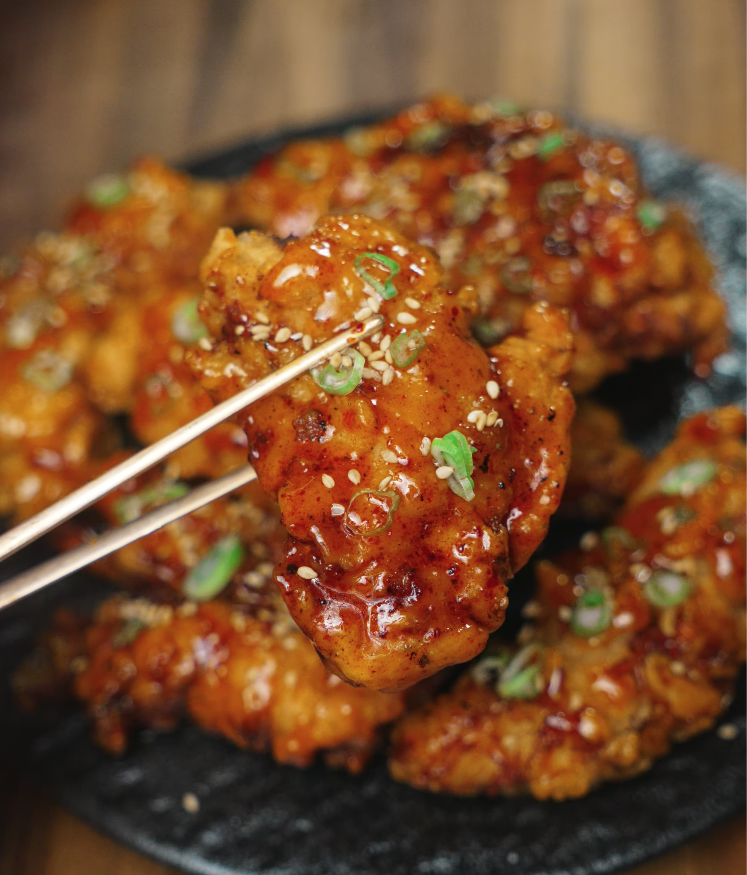 Close-up of a glistening hot honey chicken tender held up by chopsticks, highlighting the sticky glaze, sesame seeds, and green onion garnish.