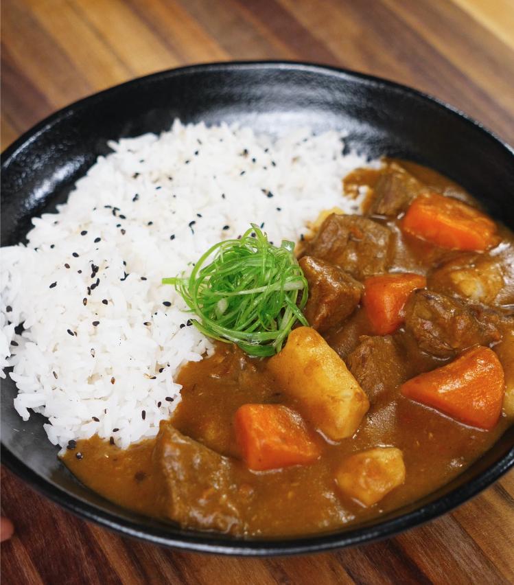 A finished bowl of Japanese beef curry served with white rice, garnished with finely shredded scallions and black sesame seeds. The rich, glossy curry features chunks of beef, carrots, and potatoes.