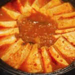 Kimchi jjigae simmering at a rapid boil with tofu slices fanned around the edges of the pot. The rich, red broth bubbles intensely, indicating deep flavor development and the perfect stage before serving.