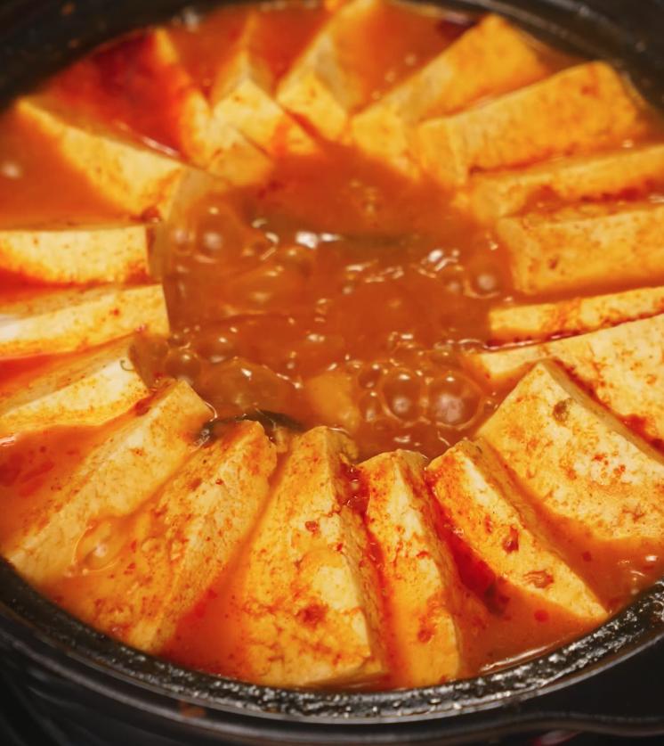 Kimchi jjigae simmering at a rapid boil with tofu slices fanned around the edges of the pot. The rich, red broth bubbles intensely, indicating deep flavor development and the perfect stage before serving.
