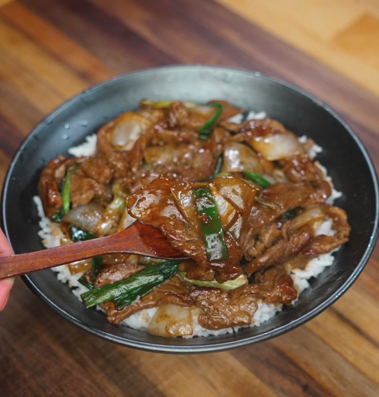 Scooping a bite of beef onion stir fry with a wooden spoon from the bowl, showcasing glossy beef, onions, and green scallions.
