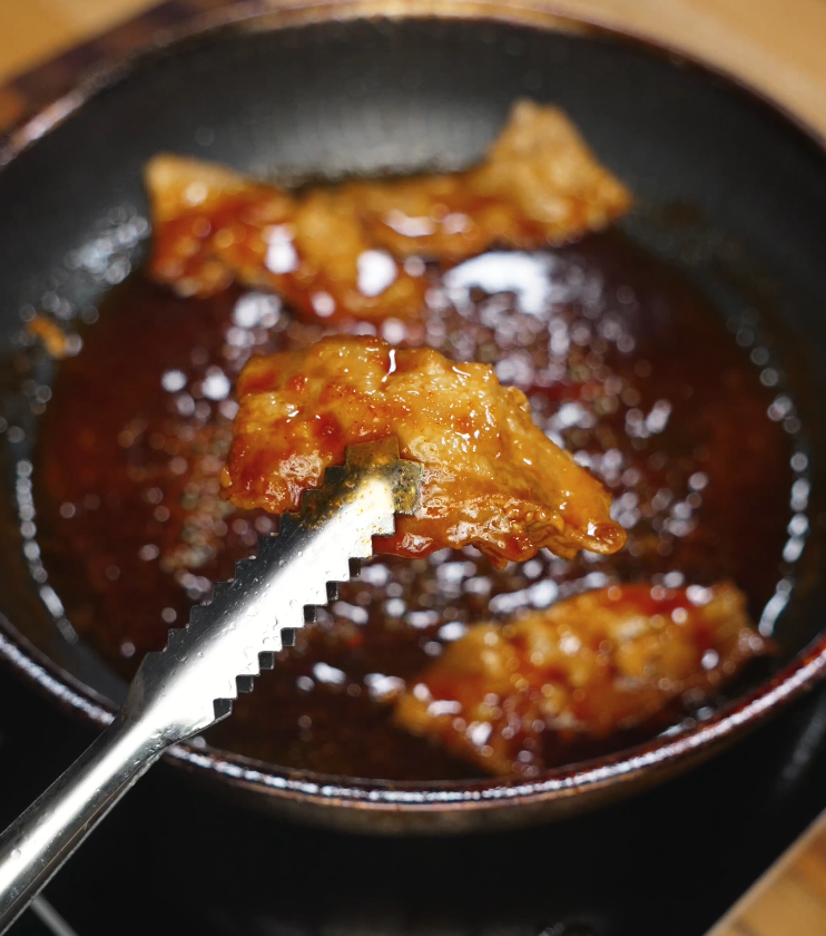 Tongs lifting a slice of fully glazed pork belly from a pan of sticky spicy sauce, showcasing the thick, glossy coating and juicy interior.