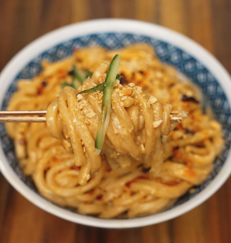 Chopsticks lifting a serving of creamy sesame noodles from the bowl, highlighting the rich sauce and garnished with fresh cucumber and crushed peanuts.