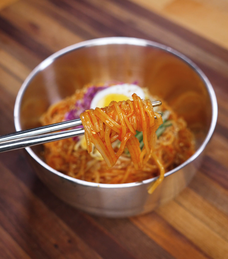 Chopsticks lift a glossy tangle of spicy jjolmyeon noodles from a metal bowl, showing their chewy texture and vibrant coating of red sauce.