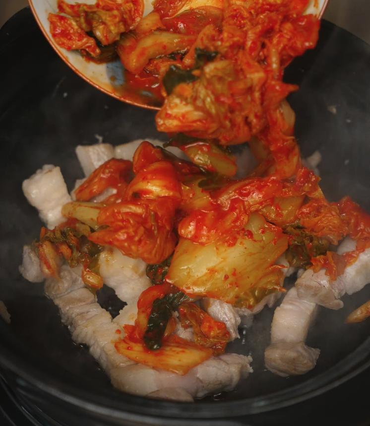 Sliced kimchi being added to seared pork belly in a pot, capturing the moment the fermented cabbage hits the heat. The combination begins to sizzle, releasing a tangy, spicy aroma that forms the base of the kimchi jjigae.
