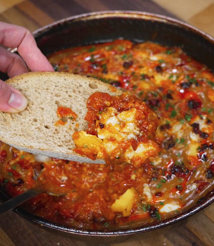 A slice of sourdough bread scooping up a rich, spicy bite of peanut chili oil shakshuka, filled with creamy egg and tomato sauce.