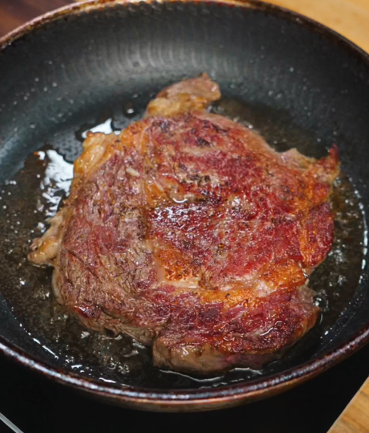 Ribeye steak sizzling in a hot pan with a deep golden crust forming for steak rice bowl.