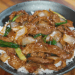 Beef and onion stir fry served over white rice in a black bowl, with glistening sauce and caramelized onions on top.