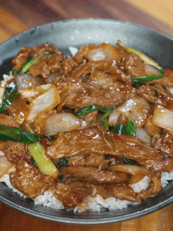 Beef and onion stir fry served over white rice in a black bowl, with glistening sauce and caramelized onions on top.