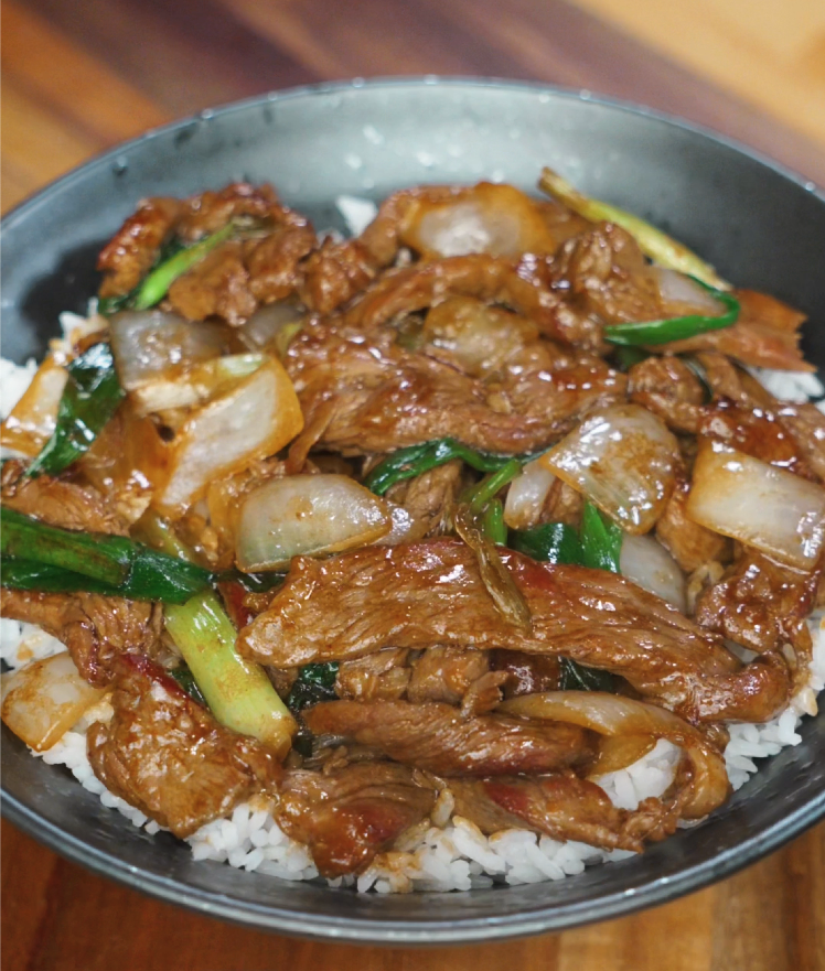 Beef and onion stir fry served over white rice in a black bowl, with glistening sauce and caramelized onions on top.