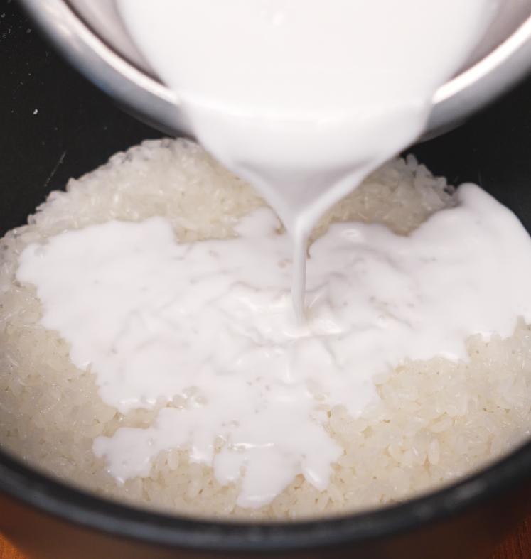 Coconut milk being poured over freshly steamed sticky rice in a pot, allowing the rice to absorb the sweet coconut flavor.