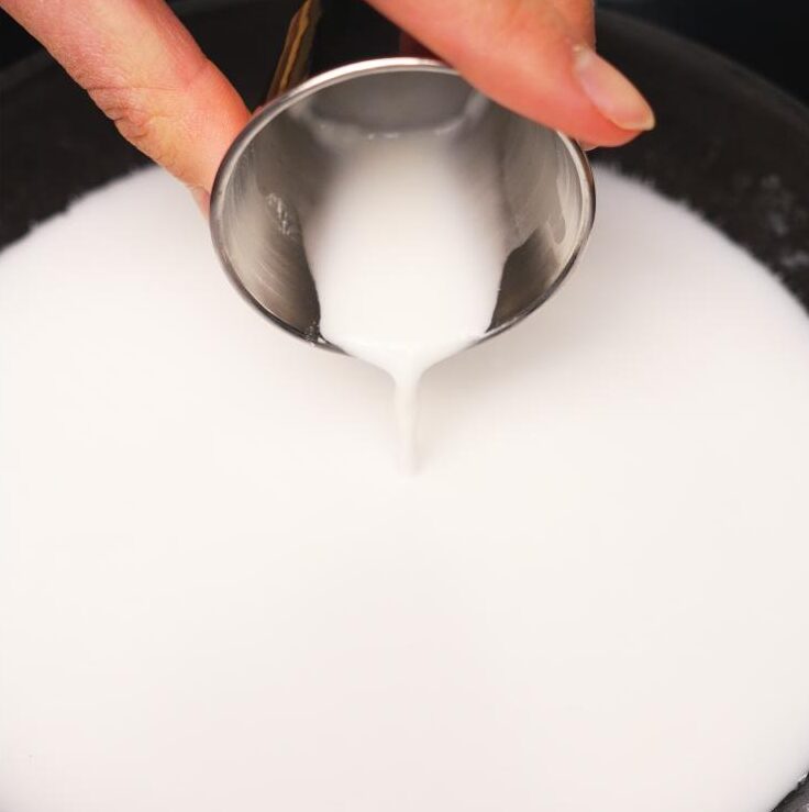Close-up of a small metal cup pouring thick coconut milk mixture into a hot pan, starting the coconut sauce for mango sticky rice.