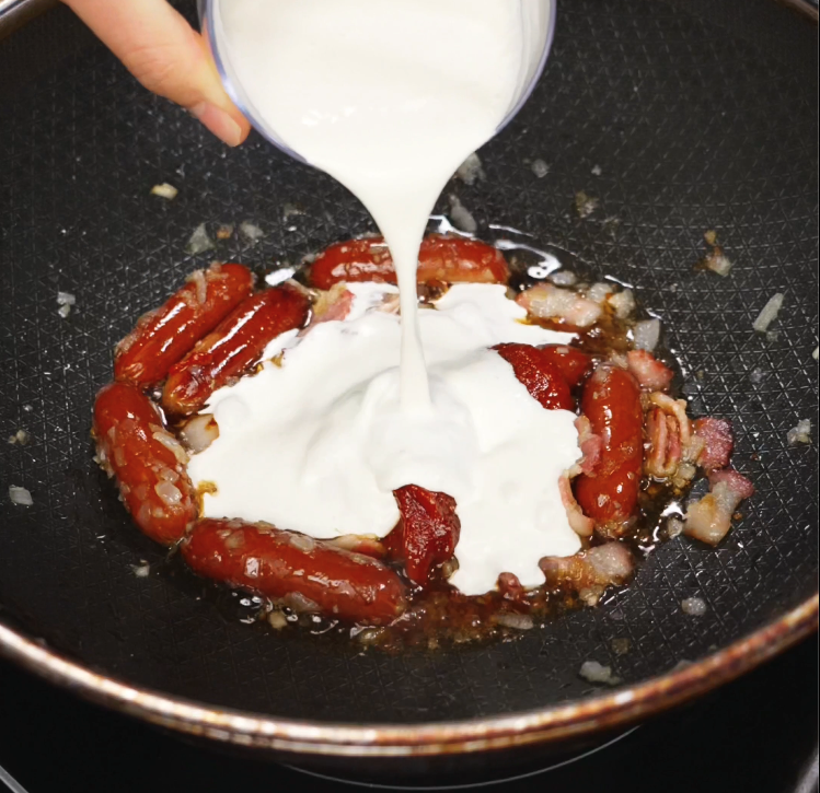 Thick cream is being poured into a pan of cooked Chinese sausage, crispy bacon, and tomato paste, forming the rich and creamy sauce for rose noodles.