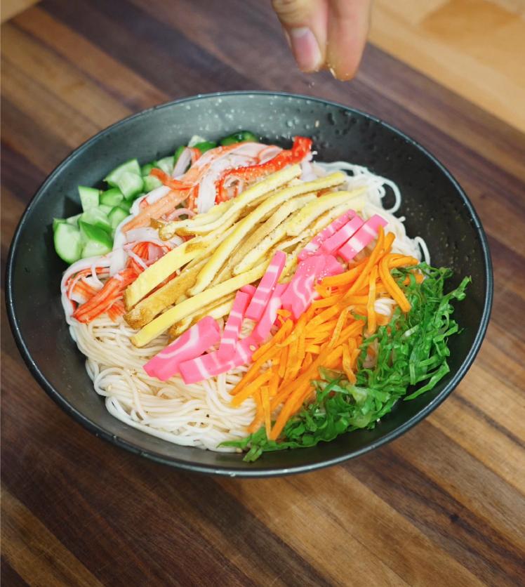A hand sprinkles sesame seeds over a colorful somen salad bowl filled with shredded egg, imitation crab, cucumber, carrots, pink fish cake, and greens arranged neatly on top of chilled somen noodles.