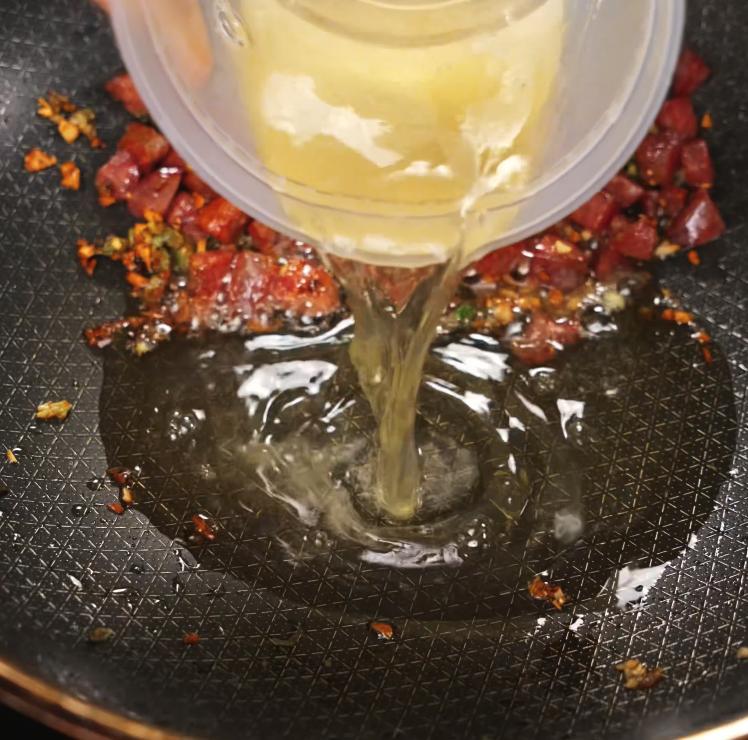 An action shot of a clear container pouring whisked eggs into a pan of sautéed lap cheong sausage, garlic, and scallions to start binding the ingredients for golden fried rice.