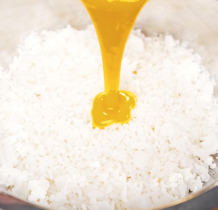 A close-up shot of vibrant golden egg yolk being poured onto a bed of fluffy day-old white rice in a metal mixing bowl, capturing the richness and color for the golden fried rice base.