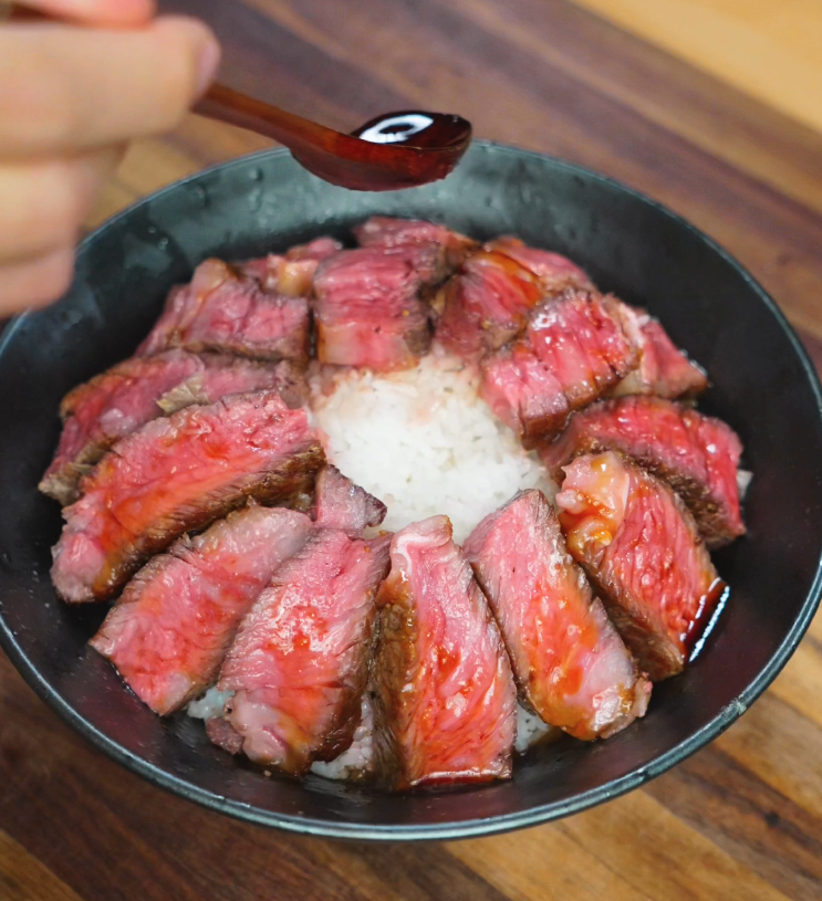 Spoon drizzling sweet-savory glaze over seared steak slices arranged on a bowl of white rice.