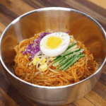 A shiny metal bowl filled with spicy jjolmyeon noodles, topped with half a boiled egg, julienned cucumber, purple cabbage, bean sprouts, and toasted sesame seeds, ready to serve.