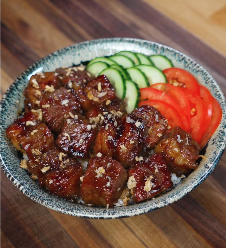 A beautifully plated bowl of shaking beef served over rice and garnished with fresh cucumber slices and tomato wedges for a vibrant, balanced presentation.