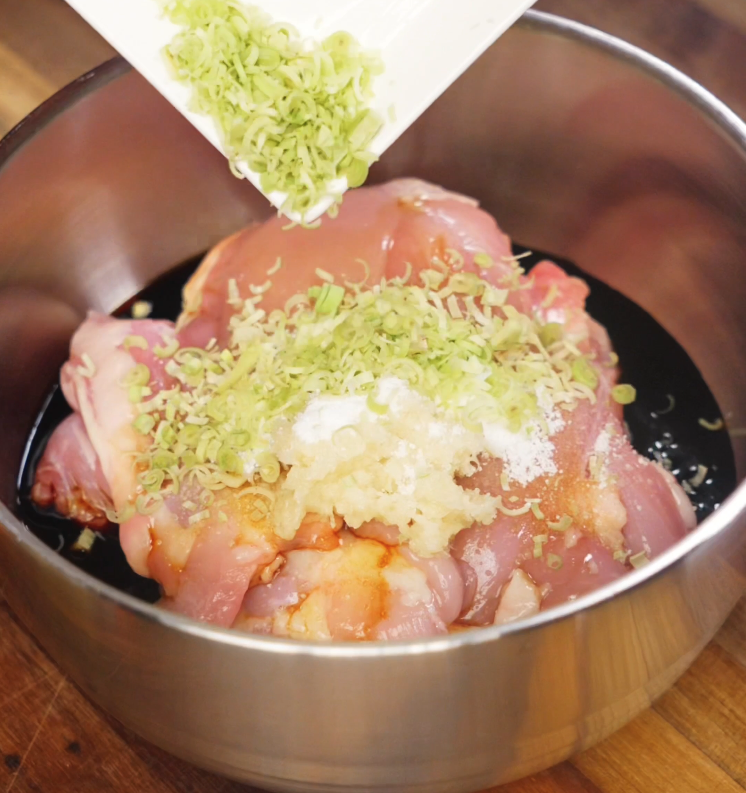 Pouring minced lemongrass over raw chicken thighs in a stainless steel bowl filled with soy sauce, sugar, garlic, and other marinade ingredients for Vietnamese lemongrass chicken.