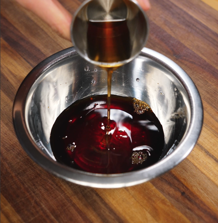 Pouring mirin from a metal measuring cup into a bowl of marinade for pork belly, with soy sauce and other seasonings visible in a stainless steel mixing bowl.