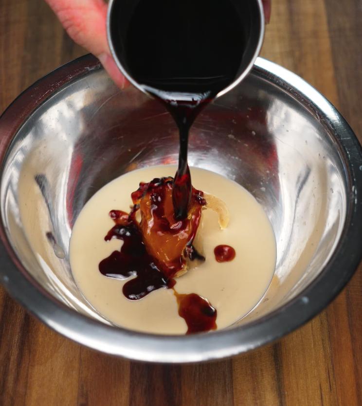 Soy sauce being poured into a mixing bowl filled with creamy sesame paste and a spoonful of peanut butter, the foundational ingredients for a rich noodle sauce.