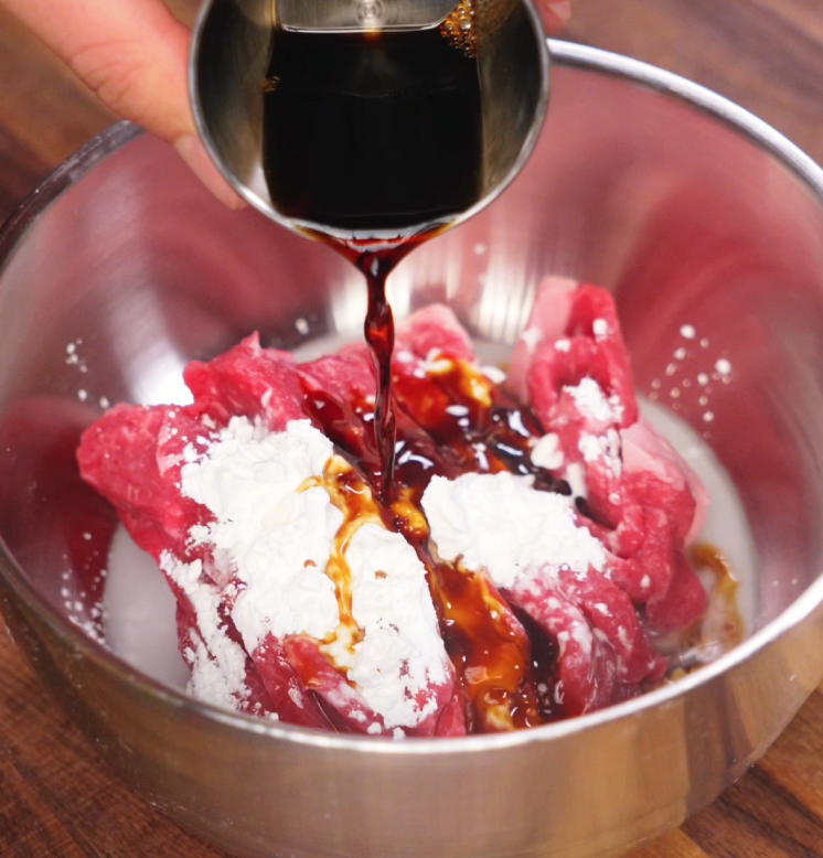 Pouring dark soy sauce marinade over sliced raw beef coated in cornstarch and milk in a metal mixing bowl, preparing for tender stir fry.