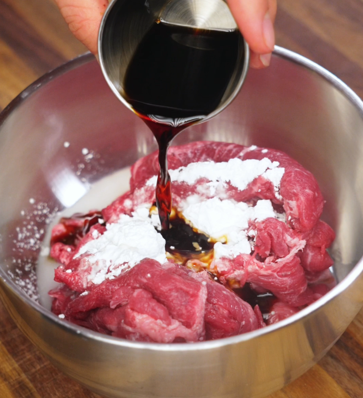 Pouring dark soy sauce onto thinly sliced beef coated with cornstarch in a stainless steel mixing bowl to start the marination for black pepper beef noodles.