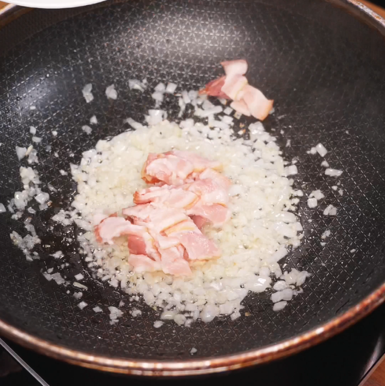 Diced onions and raw bacon pieces are being sautéed together in a nonstick pan to create the savory flavor base for rose noodles.