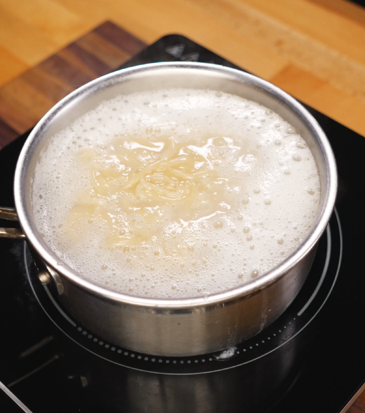 Jjolmyeon noodles are boiling rapidly in a stainless steel pot filled with starchy water on an induction cooktop, showing the cooking process to achieve their signature chewy texture.
