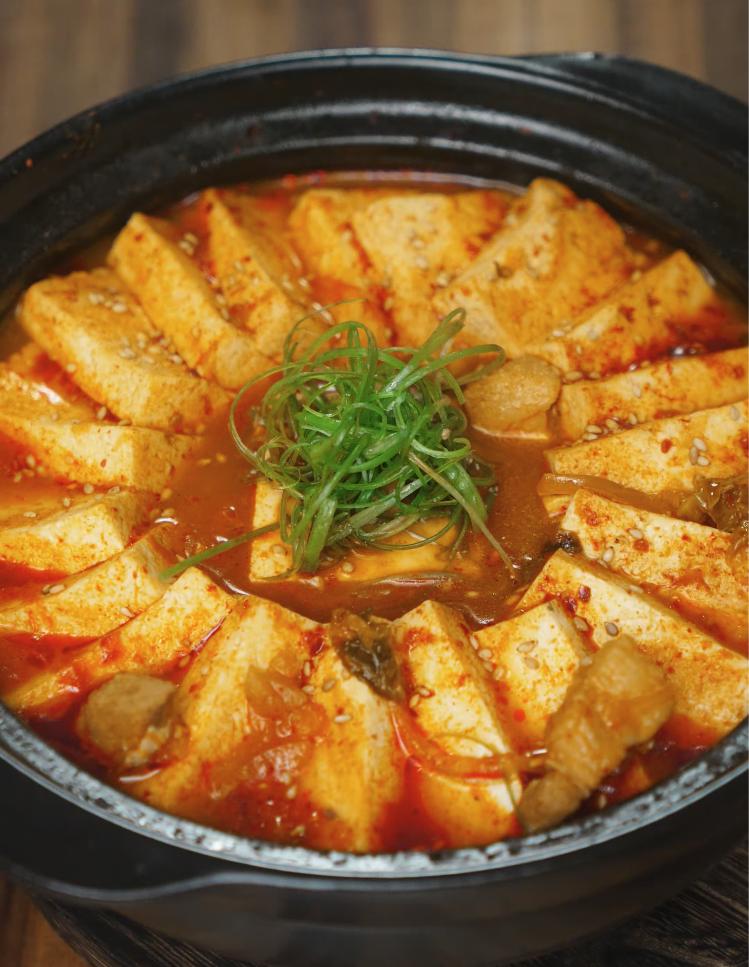 Fully cooked kimchi jjigae with tofu slices arranged in a circular pattern around the bubbling red broth, topped with thinly sliced green onions and sesame seeds. The stew is served hot in a black stone pot, showcasing its rich red hue and comforting appeal.