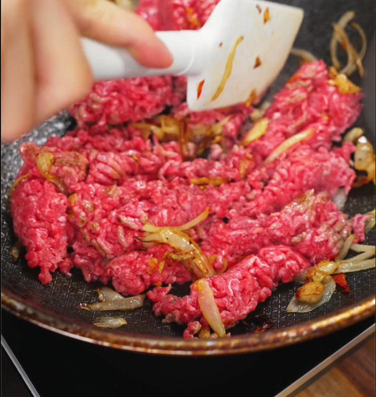 Ground beef is being browned in a nonstick skillet with sautéed onions, the base for a savory Thai pad kra pao dish.