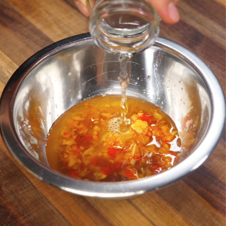 Clear rice vinegar being poured into a metal mixing bowl containing chopped garlic, red chilies, and fish sauce to make nuoc cham.