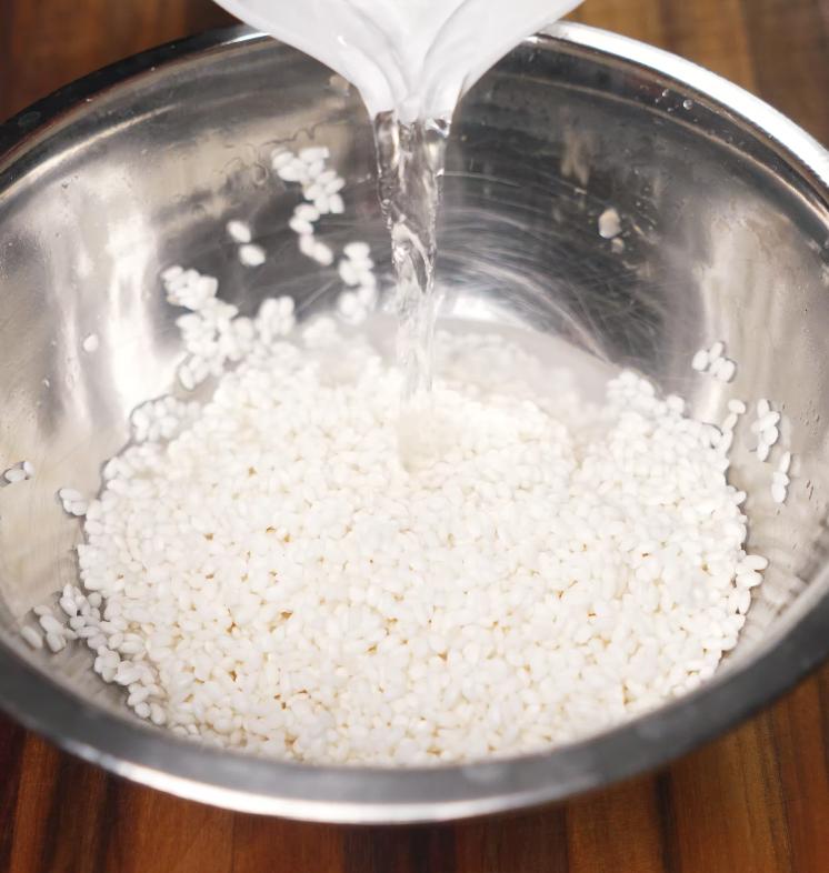 Water being poured into a stainless steel bowl filled with sticky rice, beginning the rinsing process before steaming.