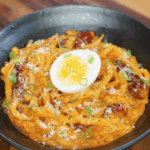 A bowl of rose noodles topped with grated cheese, chopped scallions, and a halved soft-boiled egg sits ready to serve, showcasing a rich and creamy texture.
