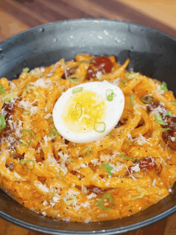 A bowl of rose noodles topped with grated cheese, chopped scallions, and a halved soft-boiled egg sits ready to serve, showcasing a rich and creamy texture.