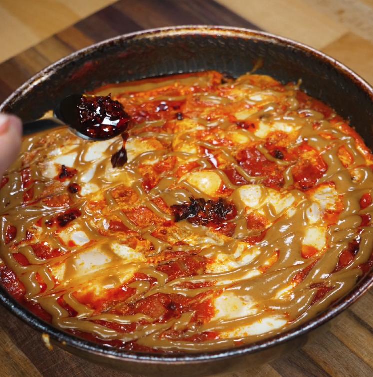A generous drizzle of chili oil and peanut butter sauce being added over poached eggs in shakshuka, creating a rich and spicy finish.