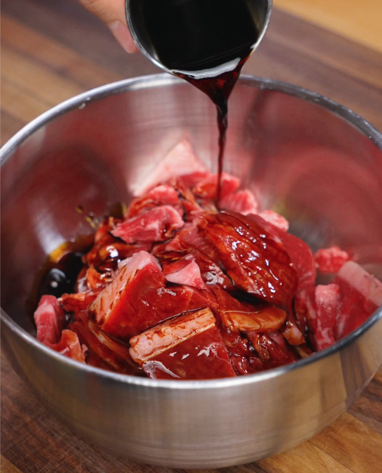 Pouring soy sauce and vinegar over raw beef slices in a metal bowl to prepare the flavorful marinade for beef tapa.