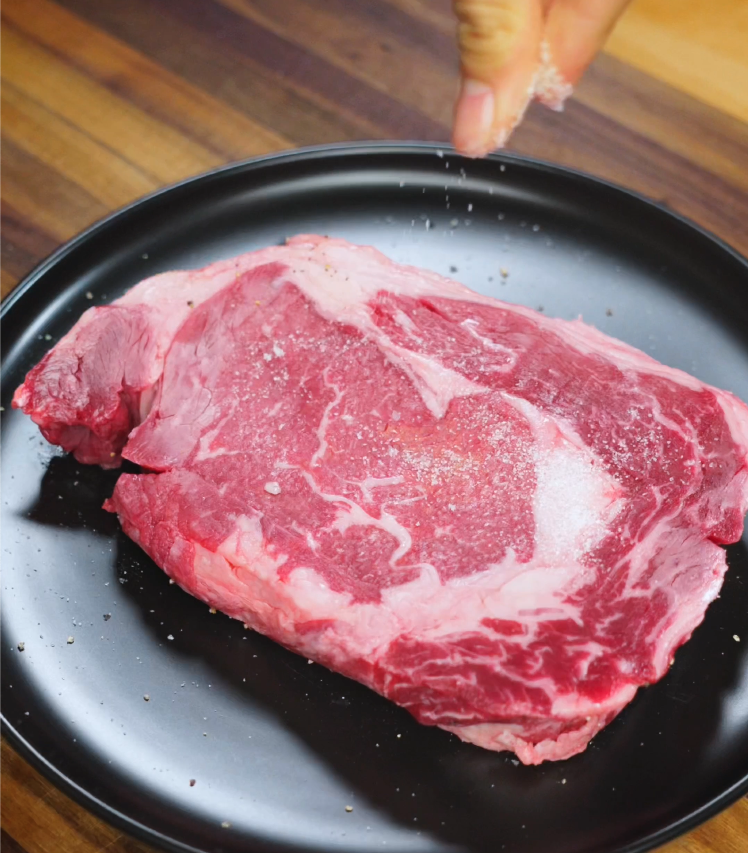 Hand sprinkling salt over a raw, marbled ribeye steak on a black plate before searing for steak rice bowl.
