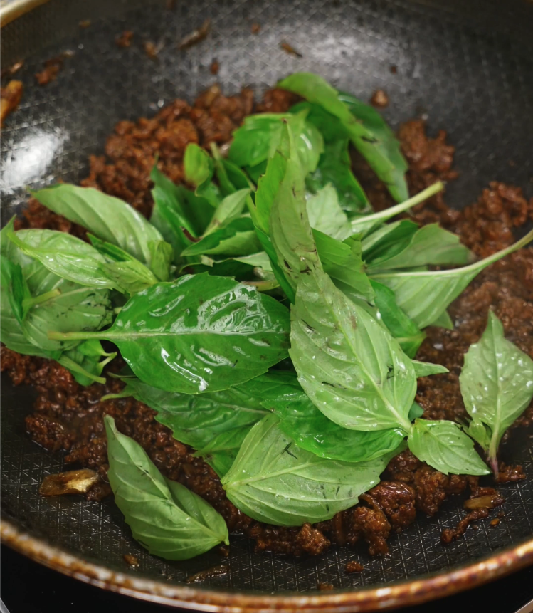 A generous handful of fresh Thai basil leaves is added to the hot skillet, releasing their aroma into the finished pad kra pao.