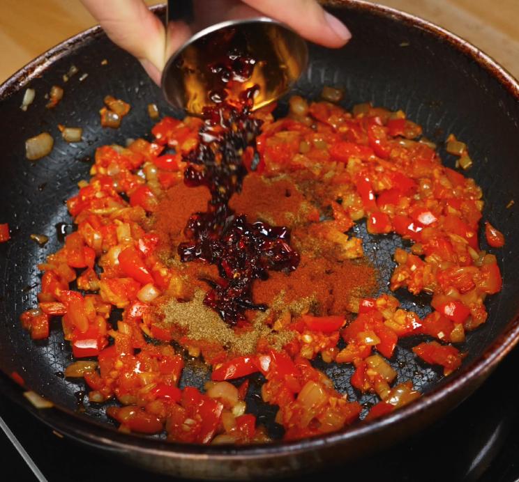 A spoonful of rich chili oil being poured into a skillet of sautéed onions, garlic, and chopped red peppers to build a savory, spicy base for peanut chili oil shakshuka.