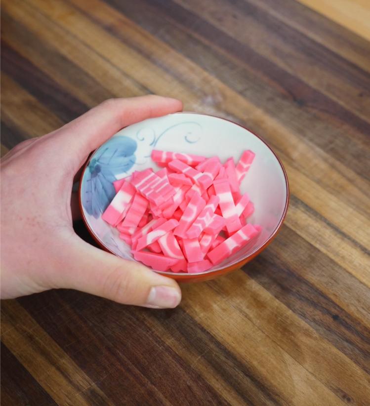 A bowl of sliced pink and white fish cake (kamaboko) being held over a wooden countertop, ready to be added to the somen salad as a colorful protein topping.