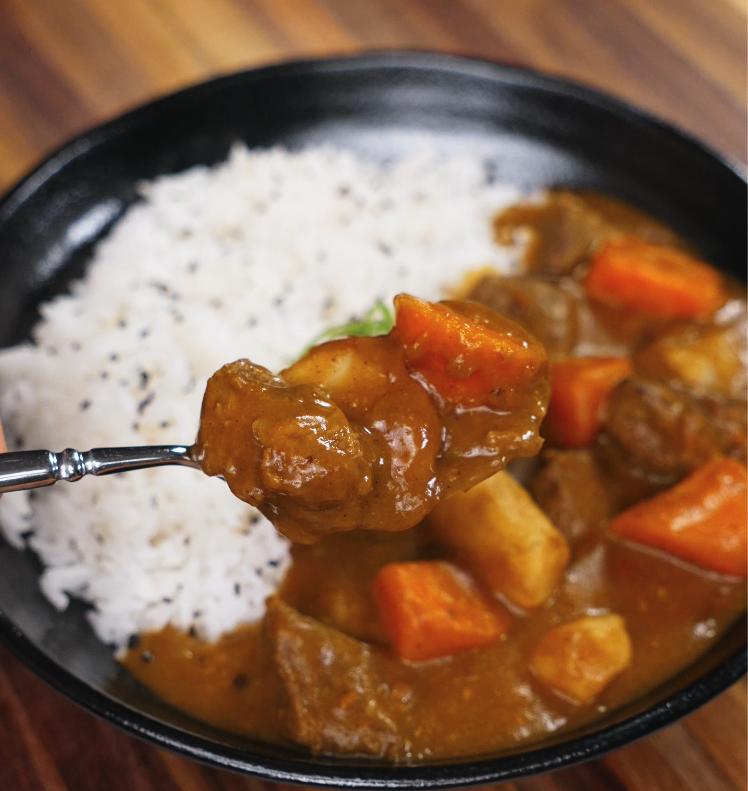 A spoonful of Japanese beef curry is lifted from the bowl, showing tender chunks of beef, carrot, and potato coated in a luscious curry sauce.