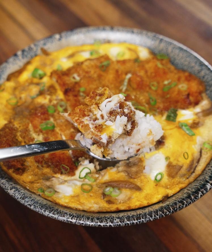 A spoonful of katsudon showing layers of crispy pork, soft egg, and white rice lifted from the bowl.
