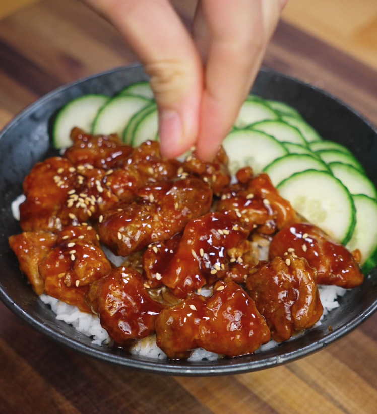 Hand sprinkling sesame seeds on top of teriyaki chicken bowl with white rice and cucumbers