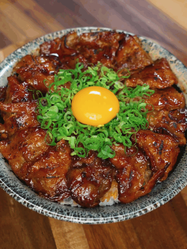 Overhead view of a sticky spicy pork belly donburi topped with finely chopped green onions and a raw egg yolk centered on top, highlighting the vibrant colors and glossy meat glaze.