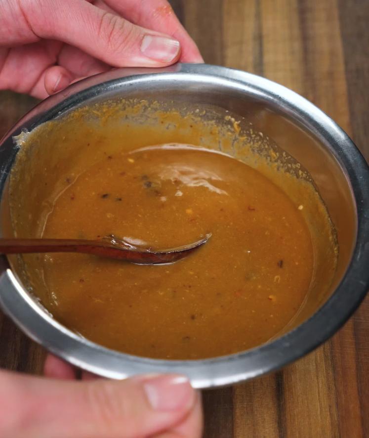 The completed spicy peanut butter sauce is shown in a metal mixing bowl, smooth and thick with a spoon resting inside, ready to be poured over the noodles.