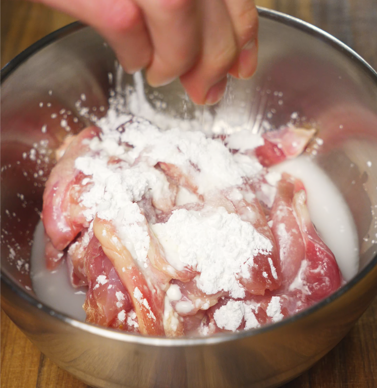Raw chicken being seasoned with cornstarch and salt in a metal mixing bowl for a tender curry texture.