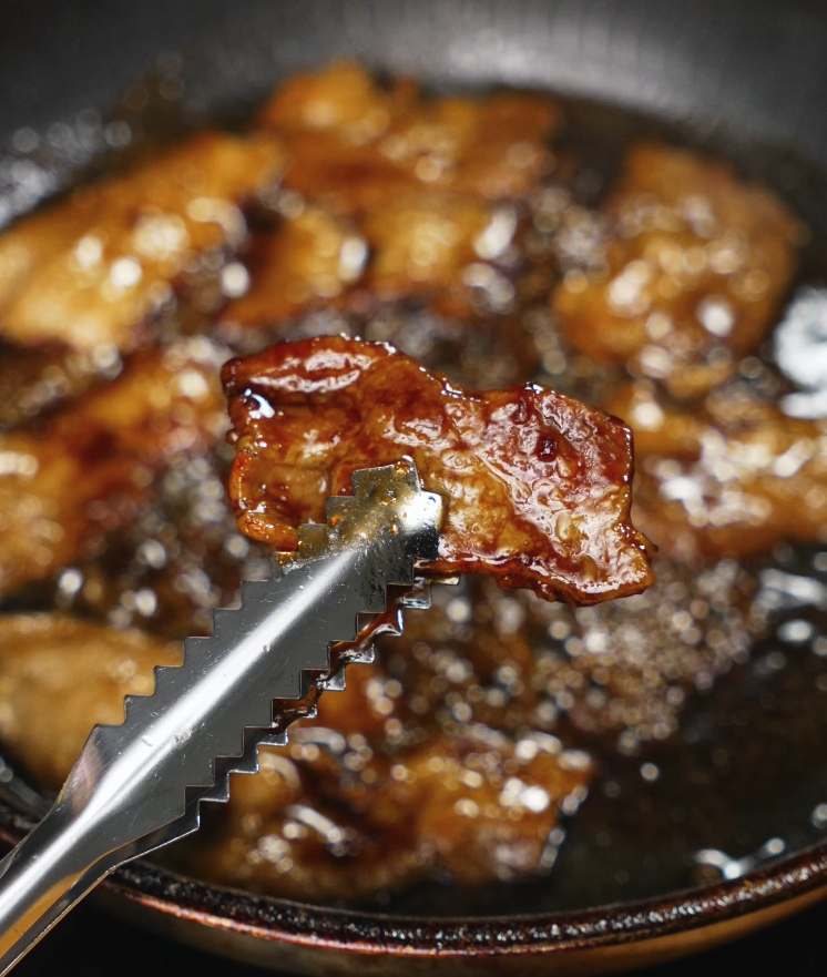 Tongs lifting a slice of fully glazed pork belly from a pan of butadon sauce, showcasing the thick, glossy coating and juicy interior.