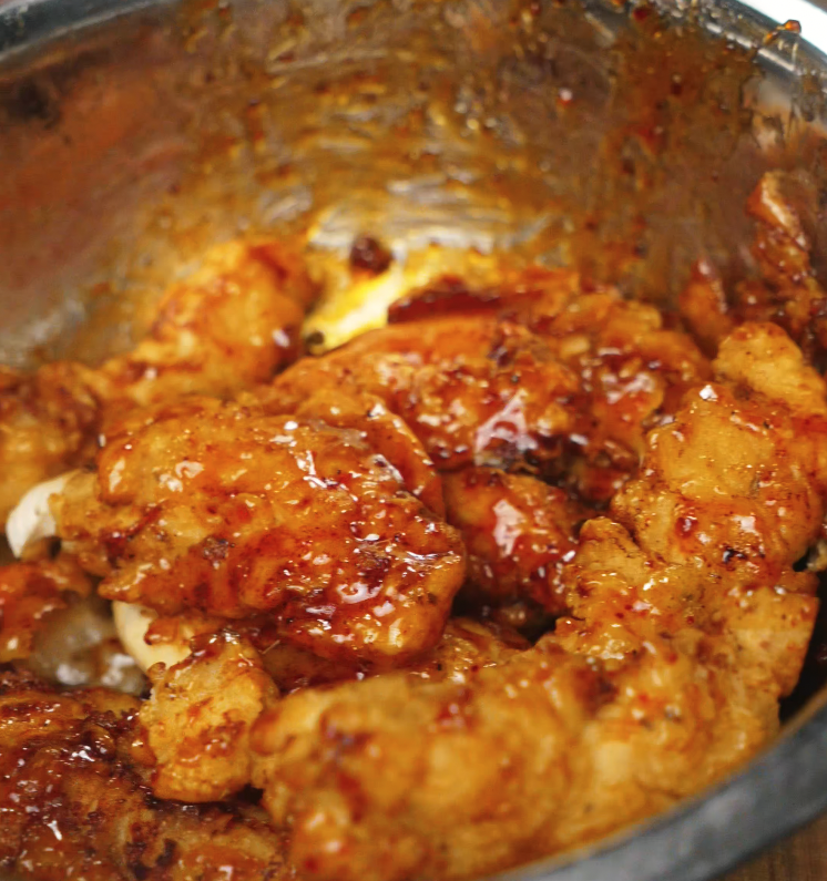 Freshly fried chicken tenders being tossed in a thick, sticky hot honey sauce inside a large stainless steel mixing bowl.